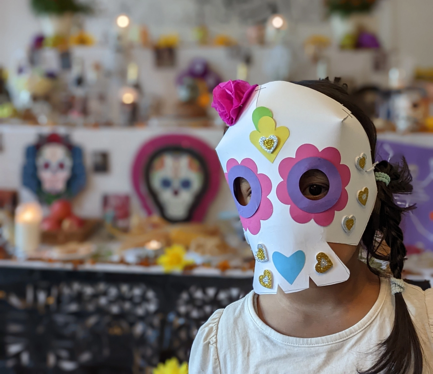 A child in a hand-decorated calavera mask with flower-shaped eyes and glitter hearts, the ofrenda lit up behind her