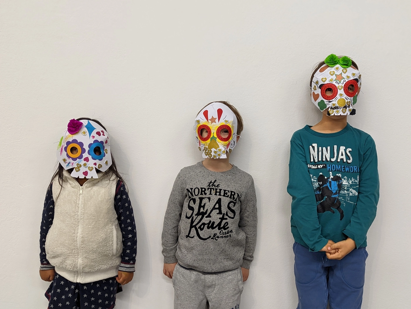 Three children lined up in the calavera masks they decorated, each one different: flowers, stars, glitter hearts