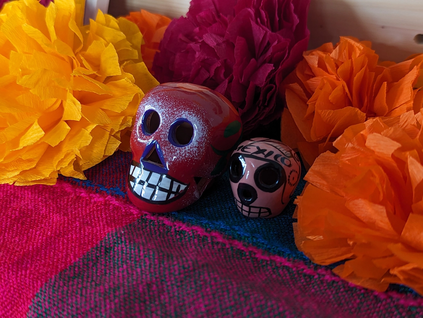 Crepe paper cempasúchil in yellow, magenta, and orange on a woven sarape, two small painted ceramic calaveras nestled between the flowers